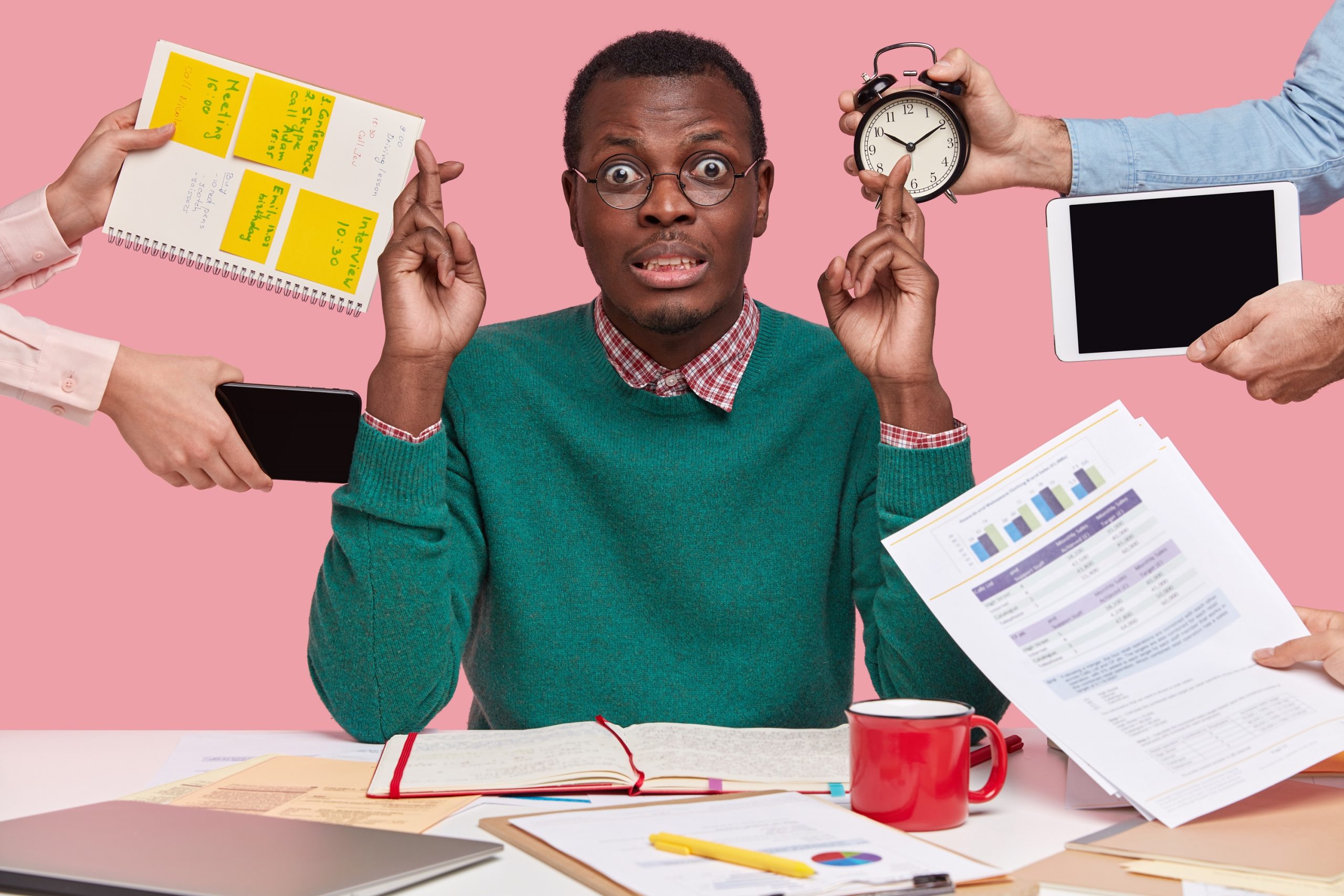 Horizontal shot of wishful dark skinned man crosses fingers, makes wish, dressed in green sweater, prepares for examination session, surrounded with alarm clock, touchpad, mobile phone, papers